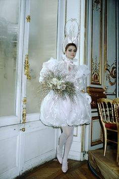 a woman in a white dress and hat is standing near a door holding a bouquet of flowers