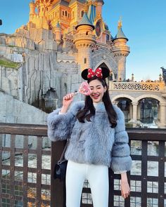 a woman standing in front of a castle wearing a mouse ears hat and fur coat