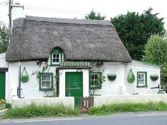 a white thatched house with green shutters on the front and side windows, next to a street