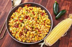 a bowl filled with corn next to two green peppers and a spoon on a wooden table