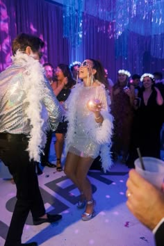 a man and woman dressed in disco wear dancing at a party with other people standing around