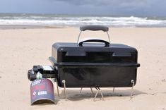 a black cooler sitting on top of a sandy beach next to the ocean with a can of beer