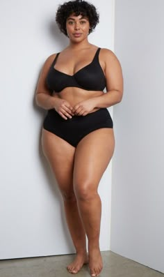 a woman in a black swimsuit leaning against a wall with her hands on her hips