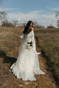 a woman in a white wedding dress walking down a dirt road with her arms around her neck