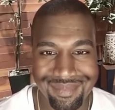 a close up of a person wearing a white shirt and smiling at the camera with plants in the background