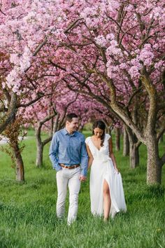 a man and woman are walking through the grass in front of trees with pink flowers