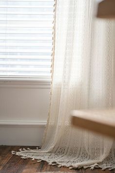 a wooden bench sitting next to a window covered in white lace curtaines with fringe trim