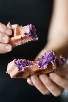 two hands holding pieces of food with purple frosting on them and one hand holding the other