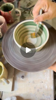 a person holding a spoon over a bowl on top of a table next to pottery