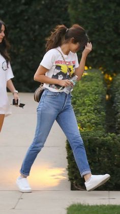 two girls walking down the sidewalk with their cell phones in hand and one is wearing white sneakers