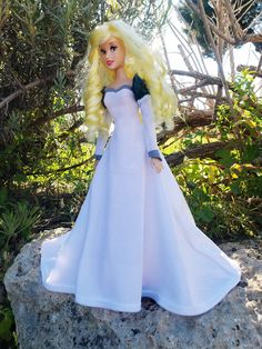 a barbie doll in a white dress standing on a rock near some bushes and trees