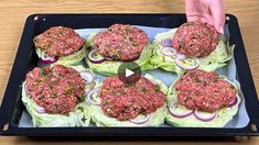 a tray filled with hamburgers and onions on top of a wooden table next to a person's hand