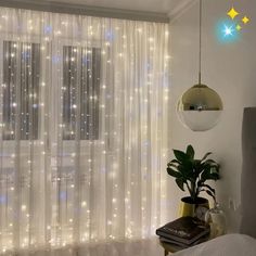 a bedroom with white curtains and lights on the window sill, next to a bed