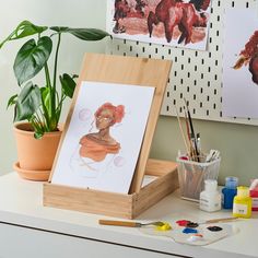 an easel with art supplies on it next to a potted plant and paintings