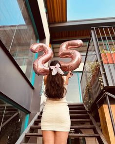 a woman standing in front of a stair case holding up two large pink number balloons