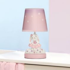 a pink lamp with a white rabbit on it sitting on a table next to a bed