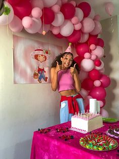 a woman standing in front of a table with cake and balloons on the wall behind her