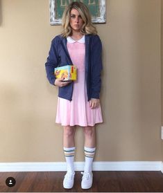 a woman in a pink dress and blue jacket is holding a box while standing against a wall