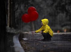 a little boy in yellow jacket kneeling down next to red heart shaped balloons on street