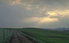 the sun shines through clouds over a green field with a dirt road in the foreground