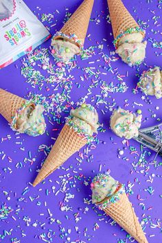 four ice cream cones with sprinkles on a purple surface next to a spoon