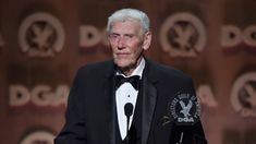 an older man in a tuxedo and bow tie giving a speech at the end of a stage