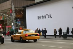 a taxi cab driving down a street next to a tall white wall with the words dear new york on it