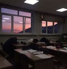 several people sleeping on desks in front of large windows at dusk with the sun going down