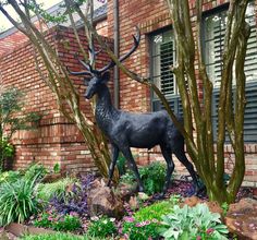 a statue of a deer in front of a brick building with trees and flowers around it