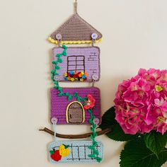 a crocheted house hanging on a wall next to a pink flower