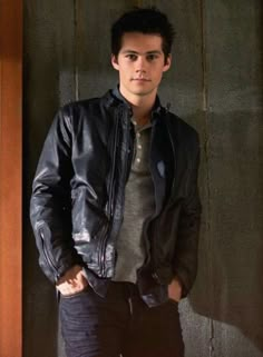 a young man standing in front of a wall wearing a black leather jacket and jeans