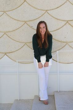 a woman standing in front of a wall wearing white pants and a green shirt with her hands on her hips