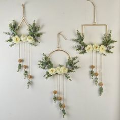 three white flowers are hanging on the wall next to some gold hoop holders with greenery