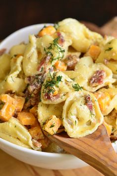 a white bowl filled with pasta and meat on top of a wooden table next to a wooden spoon