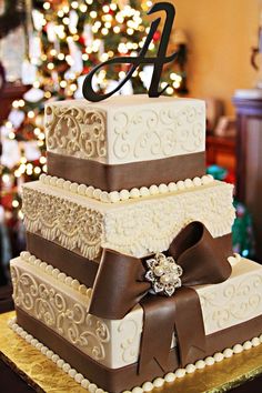 a three tiered wedding cake with brown ribbon and bow on top, sitting in front of a christmas tree