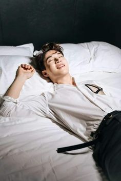 a young man laying on top of a bed next to a black bag and white sheets