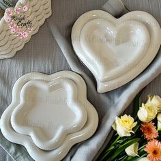 two white heart shaped plates sitting on top of a gray table cloth next to flowers