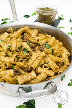 a pan filled with pasta and spinach on top of a white tablecloth next to a jar of pesto