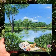 someone holding up a painting of a picnic on the river bank in front of some trees