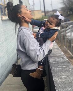 a woman holding a small child on her shoulder while standing next to a wall and looking at the camera