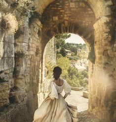 a woman in a white dress is walking through an old stone tunnel with sunlight streaming through it