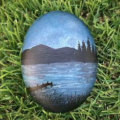 a painted rock sitting in the grass on top of a green field with trees and water