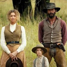 two men and a woman are standing in the grass, one is holding a hat