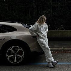 a woman leaning against the side of a car wearing an adidas sweatshirt and sweatpants