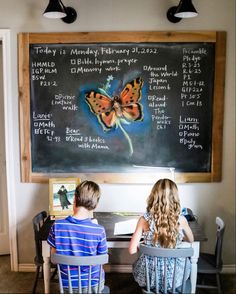 two children sitting at a table in front of a chalkboard with a butterfly on it