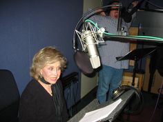 a woman sitting in front of a microphone next to a man standing behind a recording booth