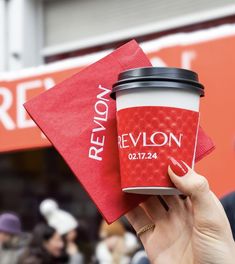 a person holding up a cup of coffee in front of a red napkin with the word revlon on it