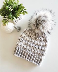 a crocheted hat with a pom - pom sits next to a potted plant