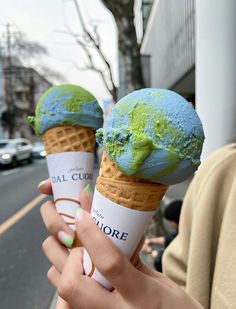 two people holding ice cream cones with green and blue toppings on them, in front of a city street