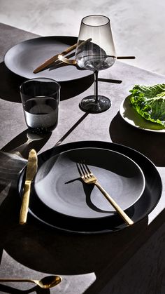 the table is set with black plates and silverware, gold utensils and green leafy greens